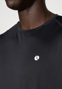 Čierne športové tričko s okrúhlym výstrihom, krátkymi rukávmi a bielym logom na hrudi, vyrobené z ľahkej, priedušnej tkaniny.