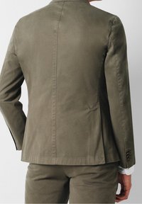 Mann in olivgrüner Anzugjacke und passender Hose, der mit dem Rücken zur Kamera gegen einen schlichten Hintergrund steht.