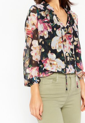 Mujer vistiendo una blusa negra transparente con grandes estampados florales en rosa y blanco y puntos dorados, combinada con pantalones verde oliva claro.