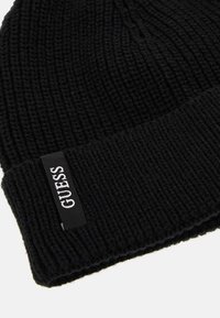 Bonnet en tricot noir avec une texture côtelée et un revers replié. Présente une petite étiquette rectangulaire avec le nom de la marque "GUESS" en lettres blanches.