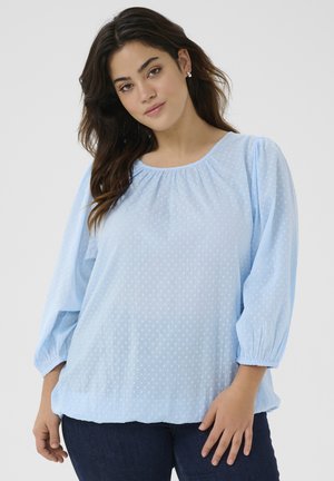 Blouse bleu clair avec des manches bouffantes et un col froncé. Présente un motif à pois texturé et une coupe décontractée au niveau de l'ourlet.