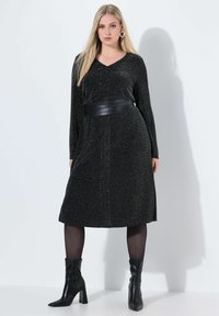 Schwarzes Strickkleid mit V-Ausschnitt, langen Ärmeln, einem Ledergürtel in der Taille und einem knielangen Saum. Getragen mit schwarzen Stiefeletten.