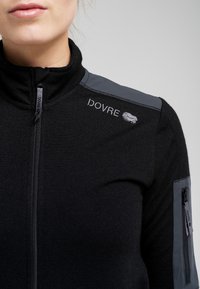 Sort fleece jakke med lynlås og grå skulderdetaljer, der har et broderet logo og en lynlåslomme på venstre arm.