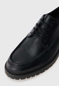 Zapatos de vestir de cuero negro con un acabado texturizado, puntera redondeada y diseño de cordones. Suela de goma negra gruesa con costuras detalladas.