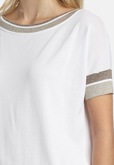 Balts īsroku krekls ar metāliskas bēša un brūnas svītras apmali apkaklē un piedurkņu galiņos, ko valkā persona no pleciem līdz vidējai rokai.