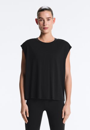 Žena s vlasy stiahnutými dozadu, oblečená v voľnom čiernom tielku bez rukávov a priliehavých čiernych nohaviciach, stojaca pred bielym pozadím.