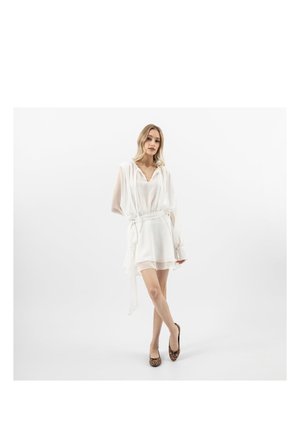 Vrouw staand met gekruiste benen, gekleed in een witte doorschijnende jurk en leopardprint ballerina's tegen een effen witte achtergrond.