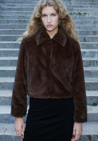 Brązowa kurtka z sztucznego futra o kroju cropped, z wykładanym kołnierzem, miękką teksturą i długimi rękawami, noszona na czarnej dopasowanej spódnicy.