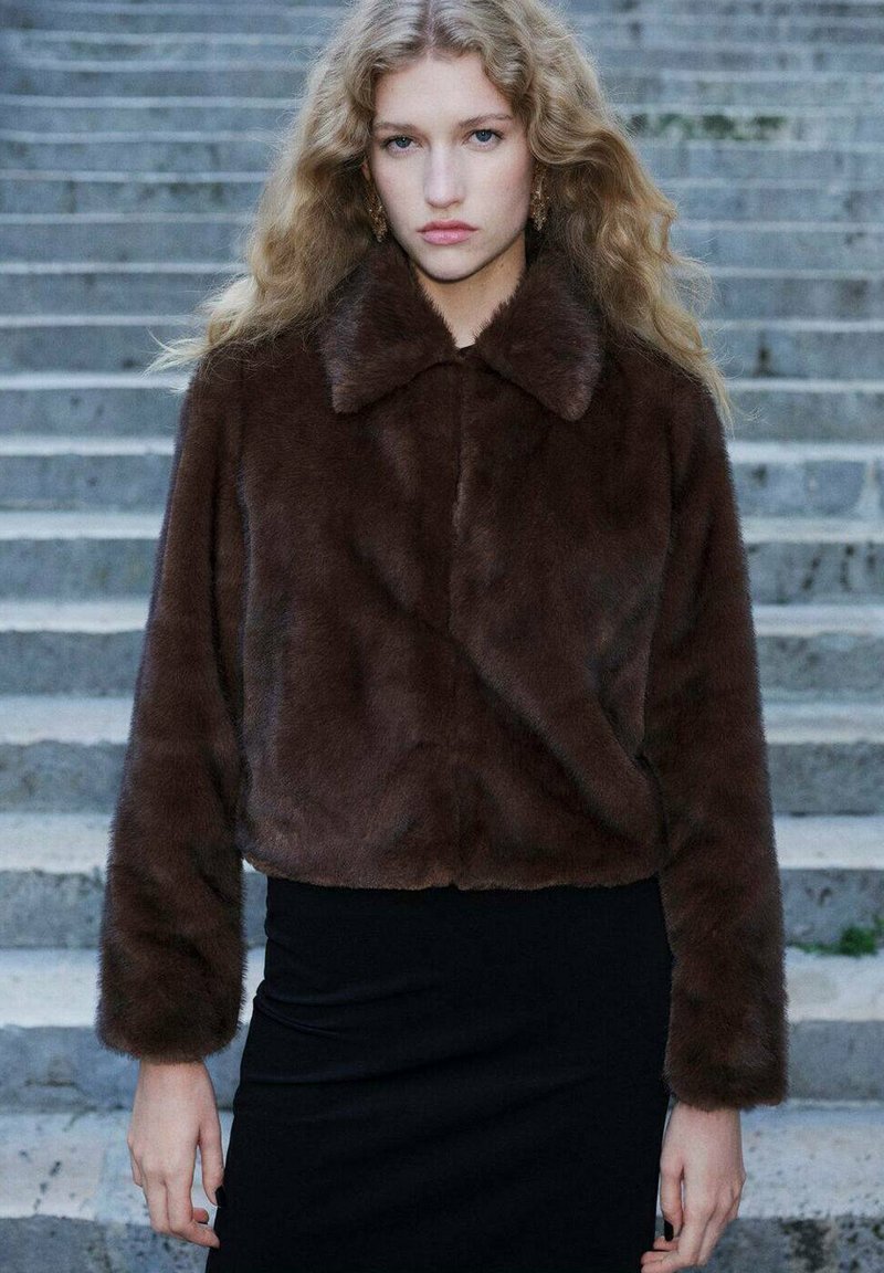 Brązowa kurtka z sztucznego futra o kroju cropped, z wykładanym kołnierzem, miękką teksturą i długimi rękawami, noszona na czarnej dopasowanej spódnicy.