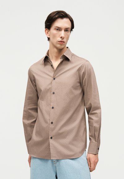 Νεαρός άνδρας που φοράει ανοιχτό καφέ πουκάμισο με μαύρα κουμπιά και ανοιχτό μπλε τζιν, στέκεται μπροστά σε απλό ανοιχτό φόντο.