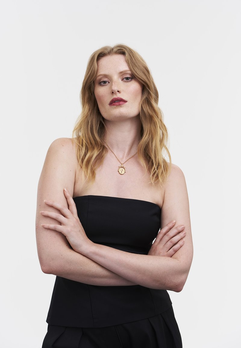 Juodas be petnešėlių viršus, dėvimas sukryžiuotomis rankomis; pasižymi lygiu paviršiumi ir struktūruotu dizainu, derinamas su subtiliu auksiniu vėriniu.