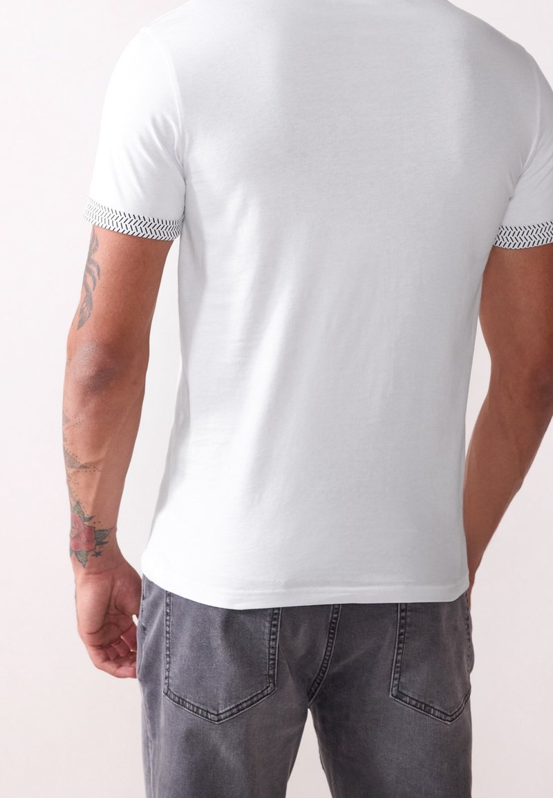 T-shirt blanc à manches courtes avec un bord noir à motifs sur les manches, associé à un pantalon en jean gris, vue de dos avec des détails de tatouage subtils.