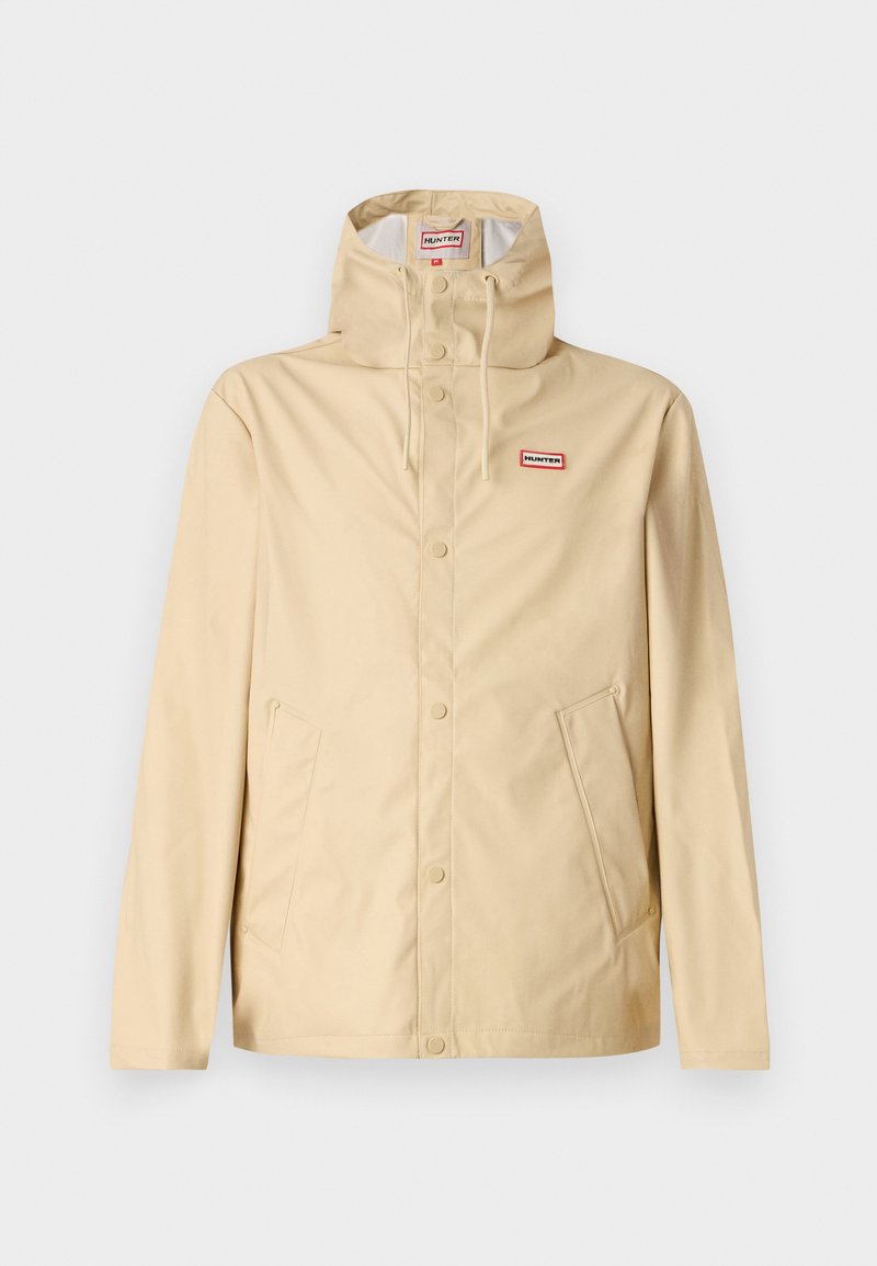 Chaqueta impermeable beige fabricada en material suave, con un cuello alto, cierre delantero abotonado y dos bolsillos laterales. Incluye capucha y logo de la marca.