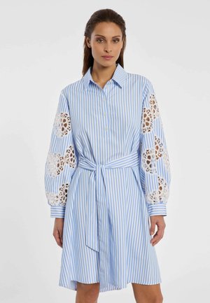 Frau trägt ein hellblau-weiß gestreiftes Kleid mit Spitzenausschnitten an den gepufften Ärmeln und einem gebundenen Taillengürtel vor einem einfarbigen Hintergrund.