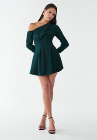 Vestido verde oscuro de un solo hombro con mangas largas, falda plisada y tela suave. Combinado con tacones negros con tiras, sobre un fondo neutro.