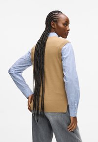 Kobieta z długimi warkoczami ma na sobie beżowy bezrękawnik na niebieską koszulę w kratę i szare dżinsy, lekko odwrócona w bok.