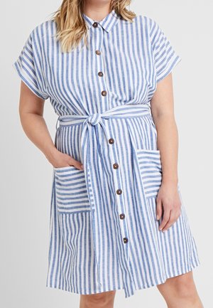 Frau trägt ein knieumspieltes weißes Kleid mit vertikalen blauen Streifen, braunen Knöpfen vorne, einem Bindegürtel und zwei aufgesetzten Taschen vorne.