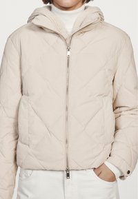 Giacca puffer beige trapuntata con chiusura a zip, cappuccio removibile e tasche laterali. Presenta dettagli in maglia a coste sul colletto.