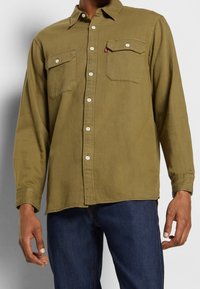 Zielona, długosleeve koszula z kołnierzykiem zapinanym na guziki, z dwiema kieszeniami na piersi i białymi guzikami, noszona na ciemnych dżinsach. Miękka tekstura materiału.