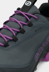Sapatilha de caminhada GORE-TEX em cinza, com atacadores roxos, detalhes pretos, um upper texturizado e o logótipo "SCARPA" em destaque.