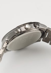 Roestvrijstalen horloge met een geborstelde afwerking, ronde kast, drie drukknoppen en een gladde, reflecterende achterkant. Verstelbare metalen band.