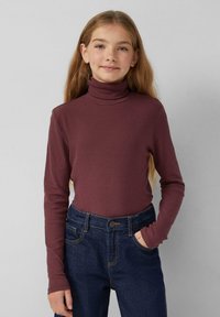 Junge Frau mit langen, hellbraunen Haaren, die einen gerippten weinroten Rollkragenpullover und dunkelblaue Jeans trägt, steht mit einer Hand in der Tasche.