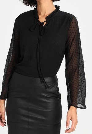 Czarna bluzka z przezroczystej, kropkowanej tkaniny, z falbankowym kołnierzykiem i wiązaniem przy szyi. Połączona z dopasowaną czarną skórzaną spódnicą.