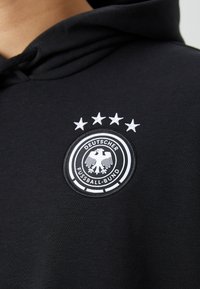 Sudadera negra con un logotipo bordado en forma de círculo, con un doble anillo y cinco estrellas en la parte superior. El logotipo incluye un águila estilizada y texto.