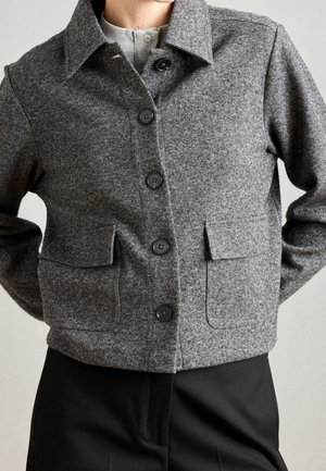 Persoon die een grijze wollen jas met knopen en klepzakken en een zwarte broek draagt, getoond van schouders tot heupen tegen een neutrale achtergrond.