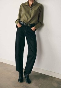 Chemise à boutons vert olive avec une poche poitrine, associée à un jean noir taille haute et des bottines noires. Ceinture avec détail de boucle métallique.