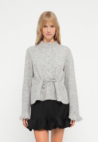 Γκρίζα πλεκτή ζακέτα με στρογγυλή λαιμόκοψη, κουμπί, κορδόνι στη μέση και μαλακή υφή. Συνδυασμένη με μια μαύρη φούστα με ρίγες.