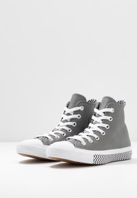 Baskets montantes en cuir gris avec des lacets blancs, des embouts en caoutchouc et des semelles noires et blanches à motif en zigzag.