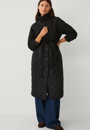 Woman wearing a long black quilted coat with a high collar and belt, paired with wide-leg blue jeans and brown shoes, standing against plain background.