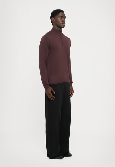 Pull rouge bordeaux avec un col zippé mi-hauteur, manches longues et poignets côtelés, associé à un pantalon large noir et des chaussures noires.