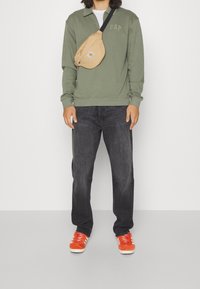 Felpa verde oliva con logo GAP, jeans neri e scarpe da ginnastica arancioni. Include una borsa a tracolla marrone chiaro con logo.