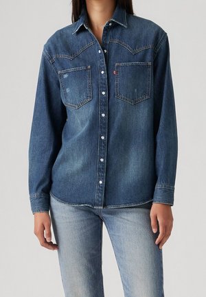 Personne portant une chemise en denim bleu foncé à manches longues avec des poches avant et un jean en denim bleu clair sur un fond uni.
