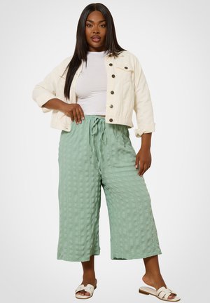 Donna che indossa una giacca corta bianca, top bianco, pantaloni larghi verde chiaro texturizzati e sandali bianchi, in piedi davanti a uno sfondo semplice.