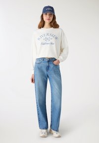 Maglietta bianca a maniche lunghe con grafica blu "Riverside", abbinata a jeans a vita alta, a gamba dritta di colore azzurro chiaro e sneakers beige. Cappellino blu.