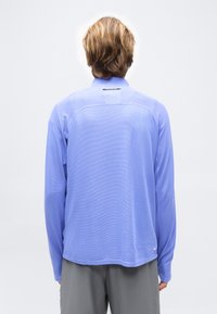 Άτομο με κοντά καστανά μαλλιά, που φοράει γαλάζιο πουκάμισο με μακριά μανίκια και γκρι παντελόνι, βλεπόμενο από πίσω σε απλό φόντο.