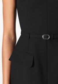Robe noire avec une silhouette ajustée, mettant en avant une taille cintrée, un détail peplum et une ceinture fine avec une boucle en argent. Tissu lisse et texturé.