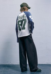 Tricou cu mâneci lungi raglan, alb și albastru, cu text verde; pantaloni negri cu croi larg; șapcă măslinie; croi lejer; fundal minimalist.