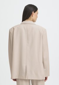 Beige tailored blazer with a relaxed fit, notched lapels, and single button closure. Features a center vent and smooth fabric texture.