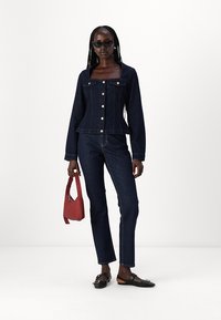 Giacca in denim scuro con bottoni dorati, abbinata a jeans coordinati. La modella tiene una borsa rossa, indossando occhiali da sole neri e scarpe.