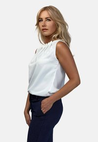 Biały bezrękawnik z marszczonym dekoltem, luźny krój. Połączony z ciemnoniebieskimi spodniami w wzory z kieszeniami. Gładka tekstura materiału.