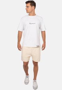 Witte katoenen t-shirt met korte raglanmouwen en zwart scriptlogo, gecombineerd met lichtbeige shorts en witte canvas sneakers.