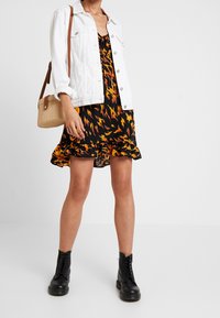 Svart klänning med orange och gul mönster, lager av volanger i nederkant, vit jeansjacka och svarta snörstövlar; brun handväska med vävd textur.