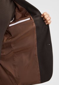 Zwarte blazer binnenkant met een bruine satijnen voering en een witte ritssluiting, zachte textuur en lichte stiksels.