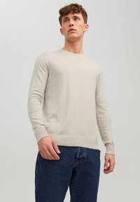 Beige knit sweater with long sleeves and ribbed cuffs, featuring a crew neckline and a slim-fit design. Worn with blue denim jeans.