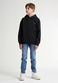 Czarna bluza z kapturem z okrągłym logo, zestawiona z jasnoniebieskimi dżinsami i białymi trampkami. Materiał wydaje się miękki i teksturowany.