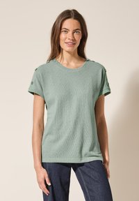 Lys grøn stribet t-shirt med korte ærmer, rund halsudskæring og dekorative knapper på skuldrene. Fremstillet af tekstureret stof.